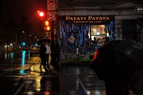 A nighttime shot of a storefront in the rain as people with umbrellas walk by.
