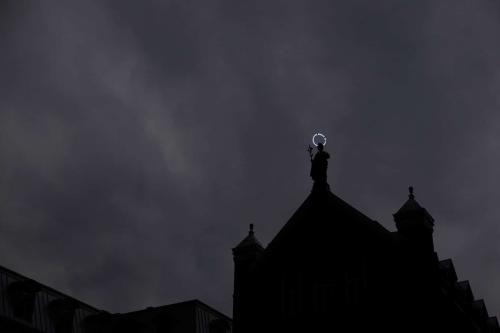 A church in silhouette, except for a Saint statue with a lit up halo, all against an gray sky