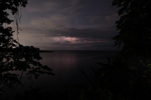 A nighttime photo of an ocean framed by foliage, with a dark sky and thunderclouds