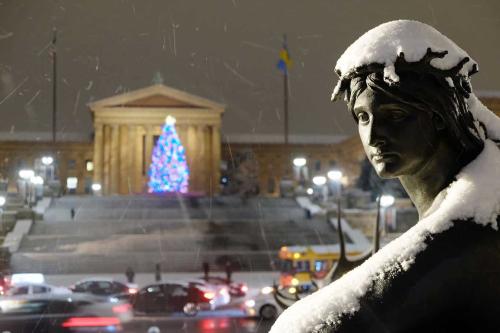 A snow-covered statue sits in the foreground as a blurred building with Christmas tree shows in the back
